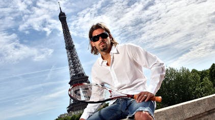 Daniel Köllerer posiert in Jeans-Shorts, weißem Hemd und Tennisschläger vor dem Eiffelturm in Paris.