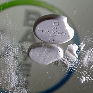 Eine Aspirin Tablette, an der eine Ecke abgebrochen ist, steht vor einem Bayer Logo auf einem Spiegel.&nbsp;