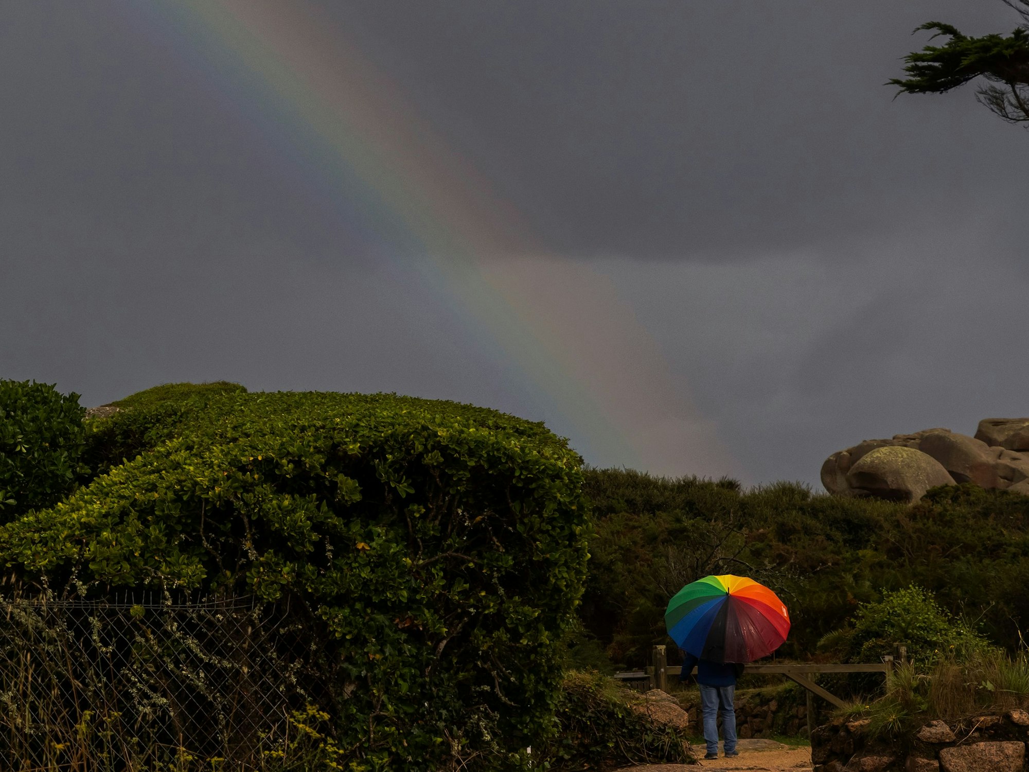 An einem grauen Himmel ist ein Regenbogen zu erkennen. Am Ende des Regenbogens steht eine Person unter einem Schirm in Regenbogenfarben.