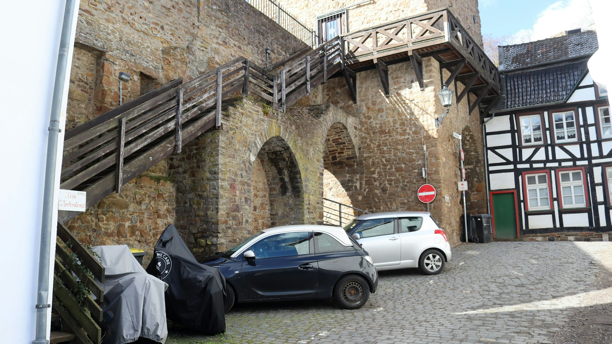 Das Bild zeigt die Holztreppe zum Wehrgang am Heisterbacher Tor aus Sicht der Werkbrücke. Vor der Treppe stehen Autos und Motorräder.