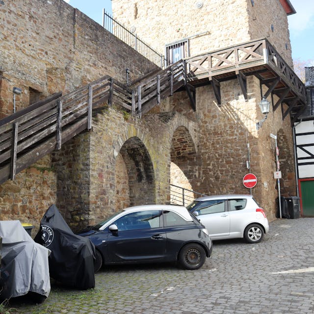 Das Bild zeigt die Holztreppe zum Wehrgang am Heisterbacher Tor aus Sicht der Werkbrücke. Vor der Treppe stehen Autos und Motorräder.