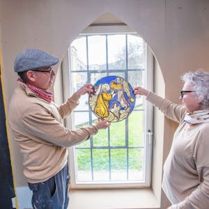 Ein Mann und eine Frau halten ein Glasmosaik in Händen, das Mosaik wird vom Tageslicht erhellt, das durch ein Fenster fällt.