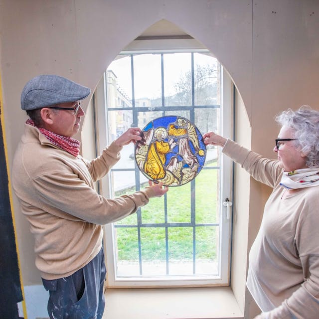 Ein Mann und eine Frau halten ein Glasmosaik in Händen, das Mosaik wird vom Tageslicht erhellt, das durch ein Fenster fällt.