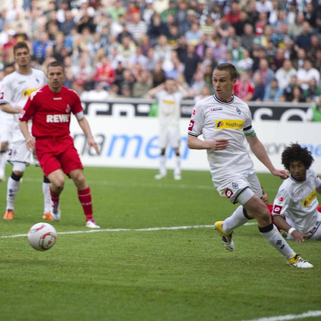 Der in rot gekleidete Stürmer des 1. FC Köln lässt zwei Gegenspieler aussteigen und erzielt ein Tor. Einer seiner Mitspieler schaut zu.