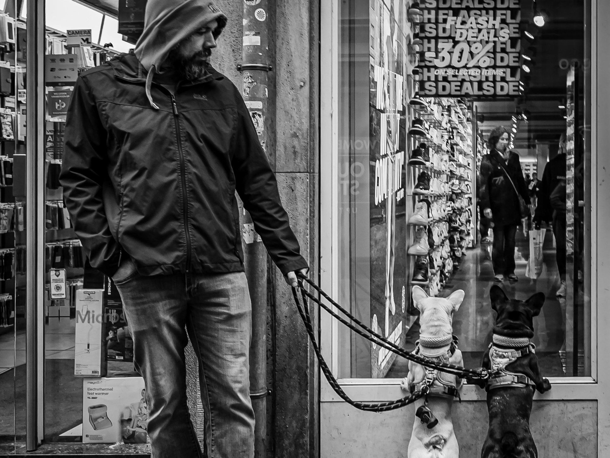 Das Schwarz-Weiß-Foto zeigt einen Mann, der zwei Hunde an der Leine führt. Diese haben die Vorderpfoten auf den Rahmen eines Schaufensters gestützt und schauen in ein Geschäft.