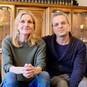 Eine Frau mit blondem Haar und ein Mann mit grauem Haar und Brille sitzen auf einem Sofa vor einer altmodischen Schrankwand.&nbsp;