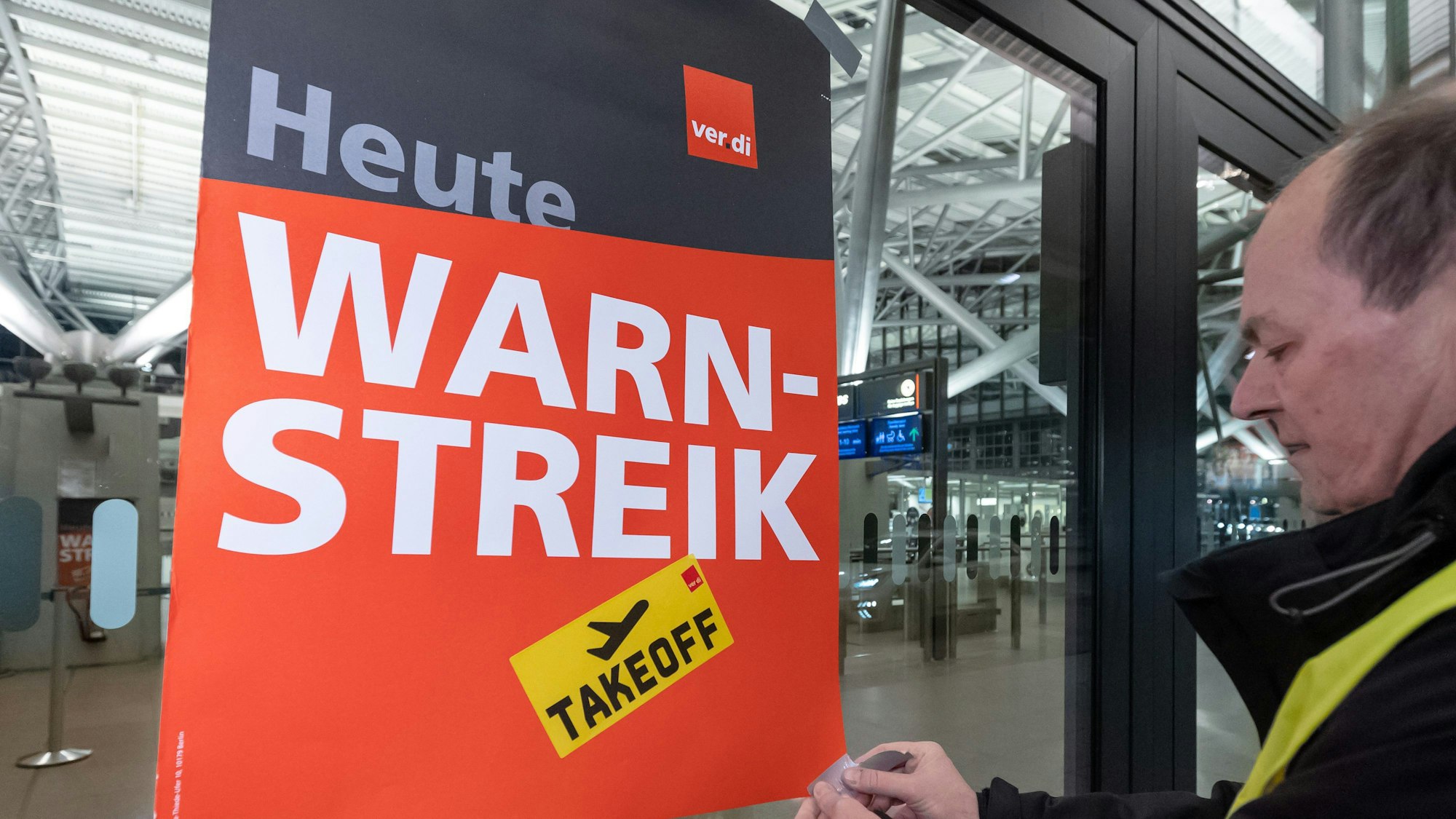Ein Verdi-Vertreter befestigt ein Plakat mit der Aufschrift «Warnstreik» an einem Terminal im Hamburger Flughafen mit Klebestreifen. Im Hintergrund ist ein leeres Flughafenterminal zu sehen.