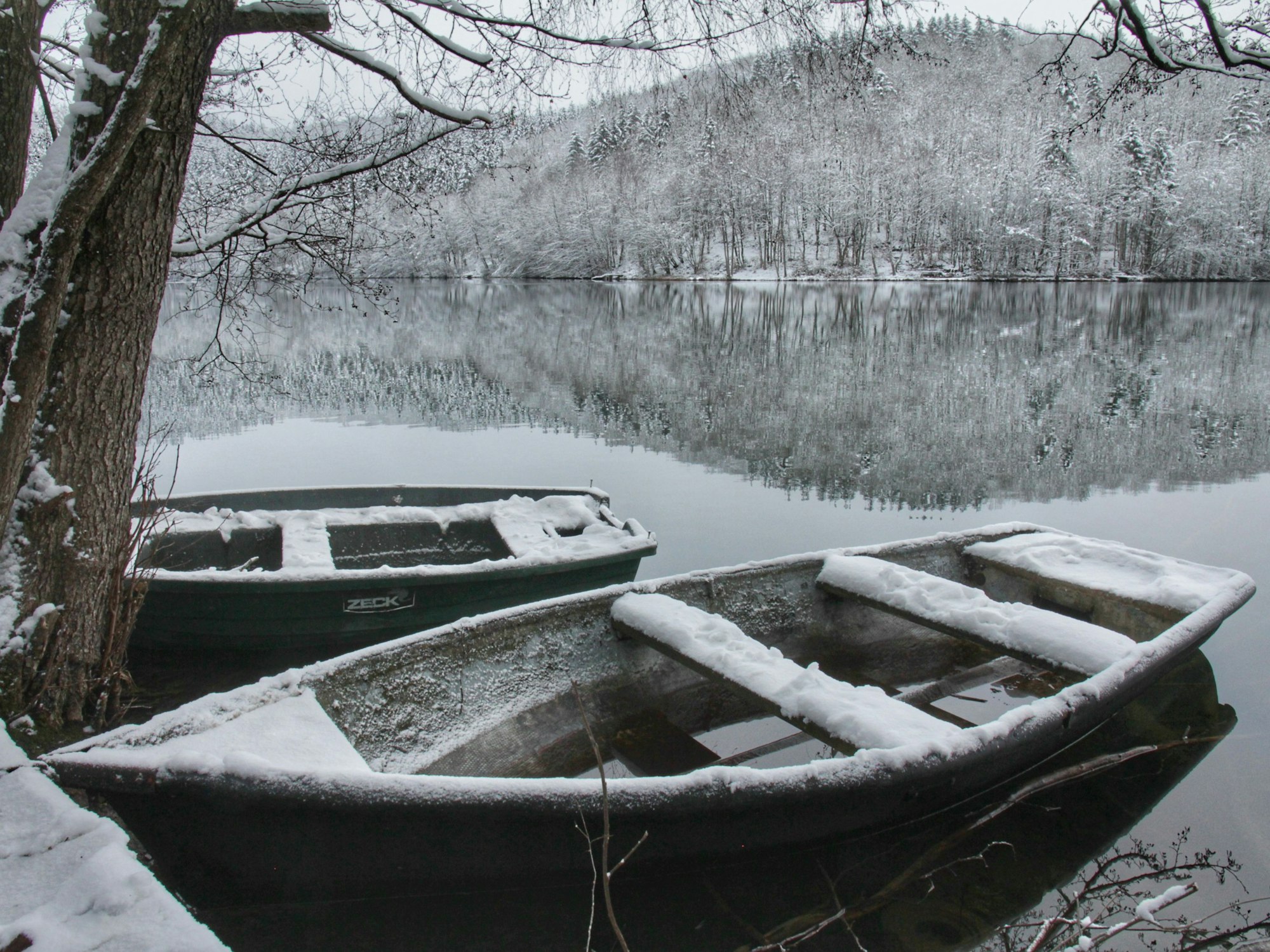 Im ruhigen Wasser des Freilinger Sees spiegelt sich die winterlich verschneite Landschaft. Im Vordergrund sind zwei Ruderboote, auf denen ebenfalls Schnee liegt.