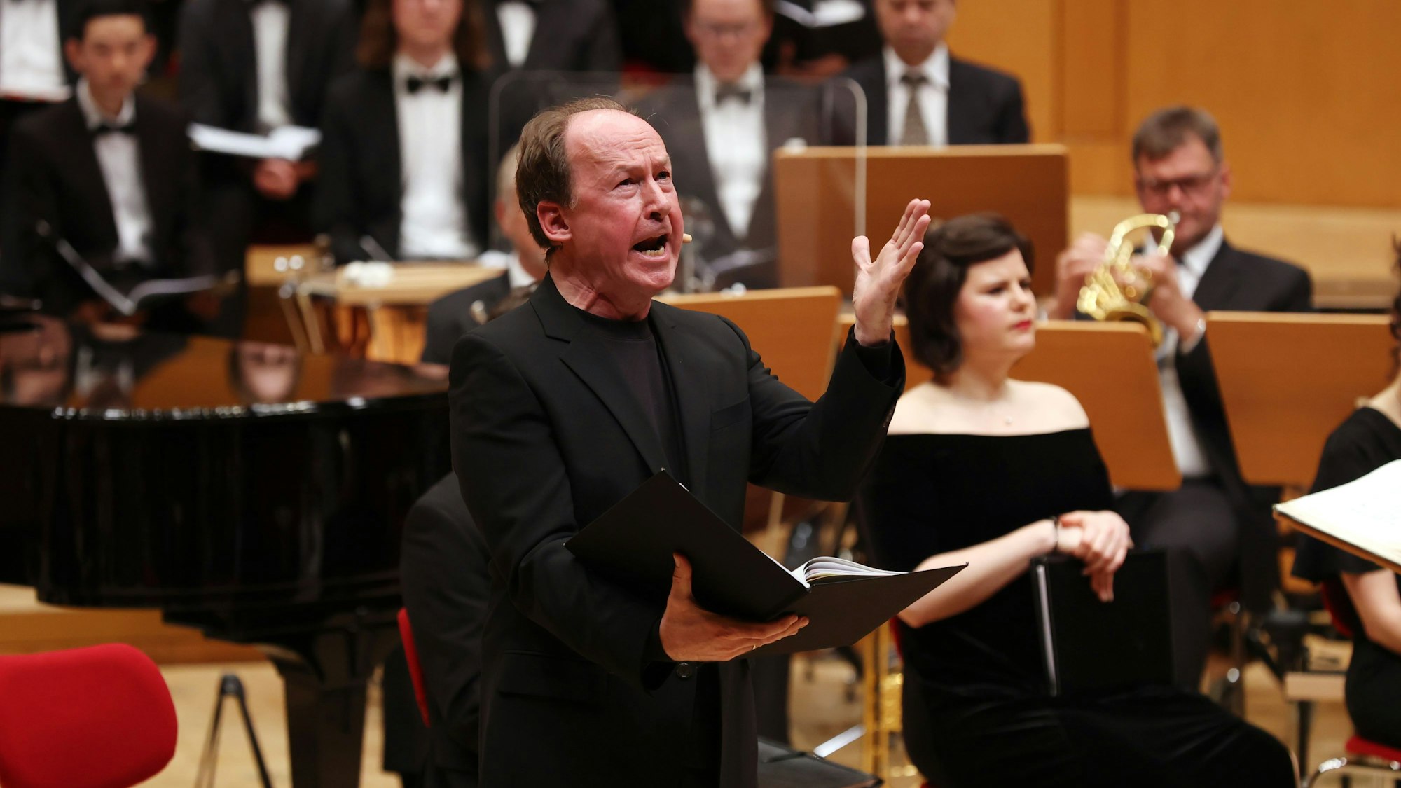 Ulrich Noethen hält eine offene Mappe in der rechten und gestikuliert mit der linken Hand. Er trägt einen schwarzen Anzug. Im Hintergrund das Orchester.