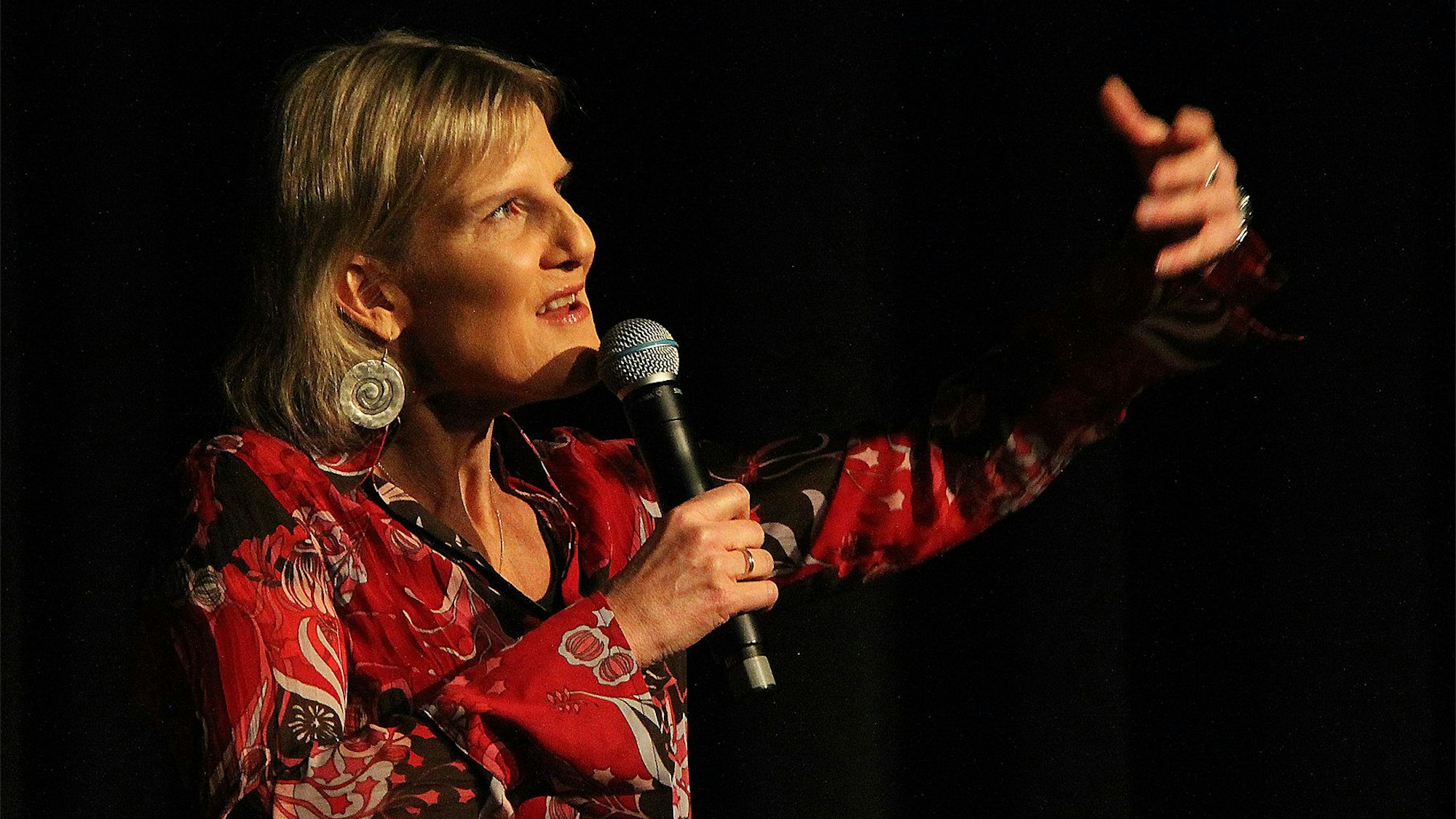 Comedienne Vera Deckers aus Köln steht in einem roten Shirt vor einem schwarzen Hintergrund auf der Bühne. Mit der linken Hand gestikuliert sie, in der rechten hält sie ein Mikrofon.
