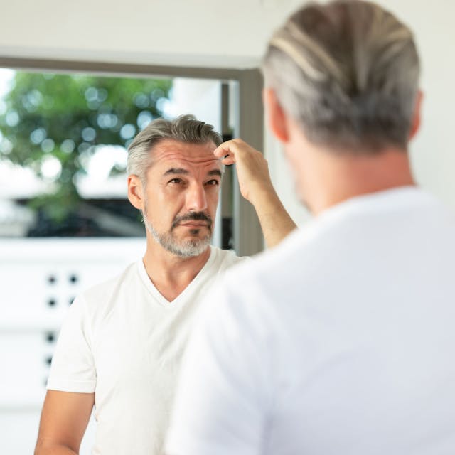 Ein Mann in weißem T-Shirt schaut in einen Spiegel und fährt sich mit der rechten Hand durch seine grauen Haare