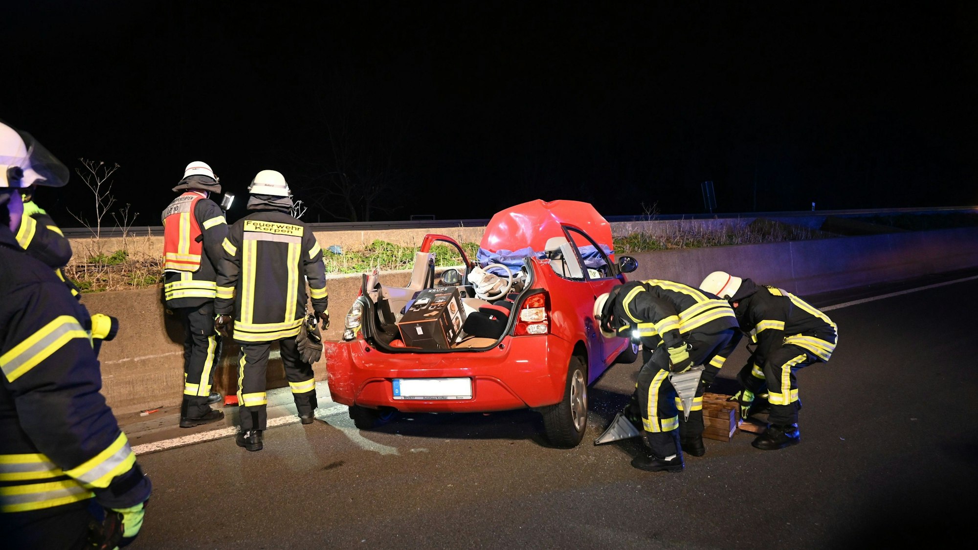 Ein rotes Auto steht an einer Betonschrammwand auf der Autobahn. Das Dach des Wagens wurde entfernt, um den Fahrer zu befreien. Feuerwehrleute befinden sich um den Wagen herum.
