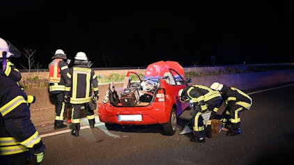 Ein rotes Auto steht an einer Betonschrammwand auf der Autobahn. Das Dach des Wagens wurde entfernt, um den Fahrer zu befreien. Feuerwehrleute befinden sich um den Wagen herum.