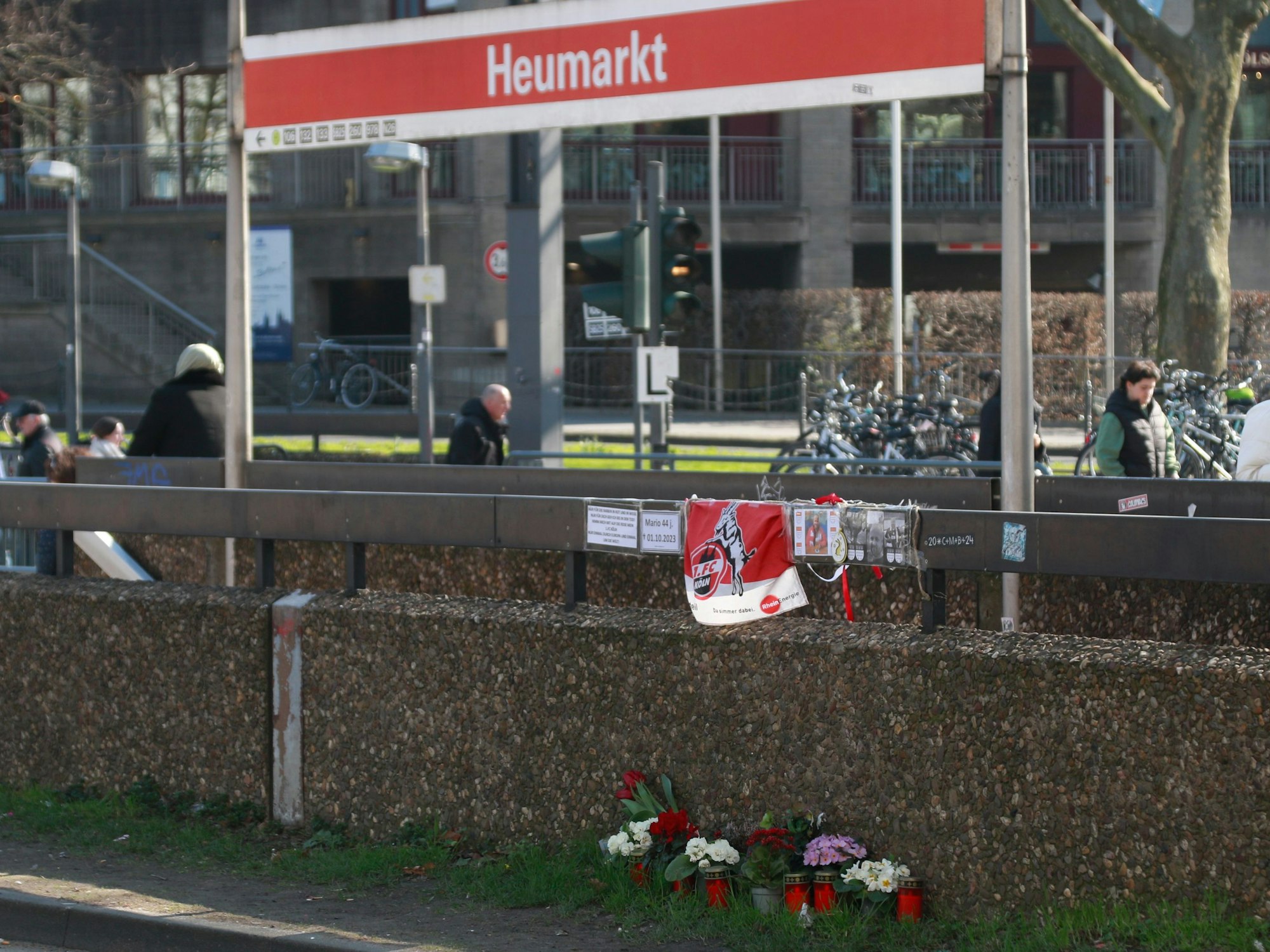 An einem Gelände hängt eine Fahne des 1. FC Köln, darunter stehen Blumen und Kerzen.