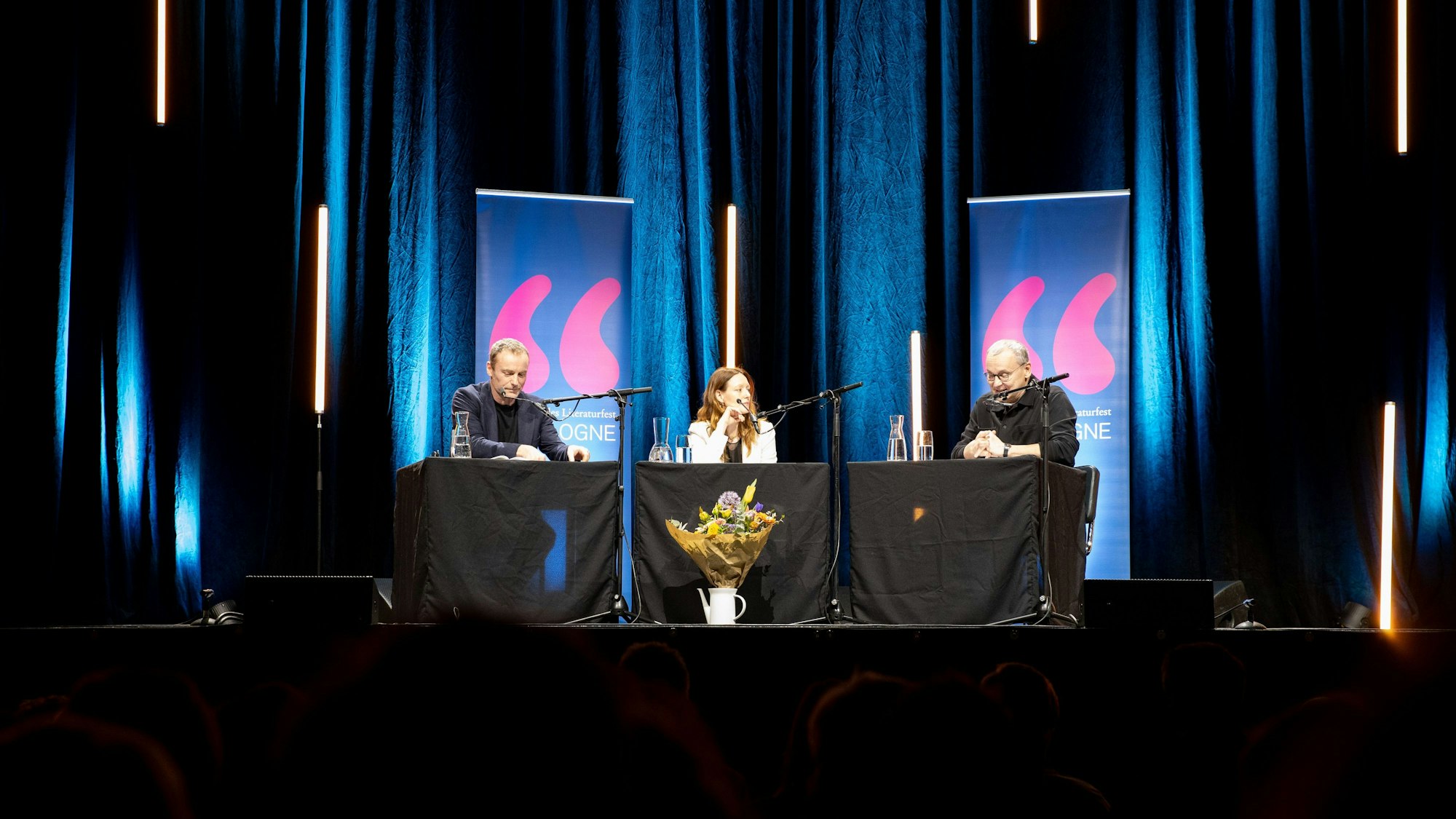 Mark Waschke, Lavinia Wilson und Knut Elstermann sitzen an drei Tischen auf der Bühne. Im Hintergrund ein blauer Vorhang und einige längliche LED-Röhren, von denen manche zu schweben scheinen-