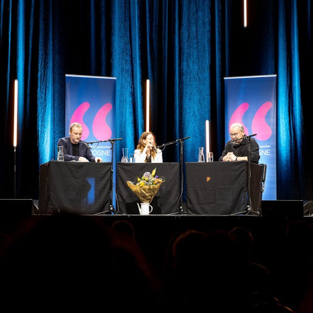 Mark Waschke, Lavinia Wilson und Knut Elstermann sitzen an drei Tischen auf der Bühne. Im Hintergrund ein blauer Vorhang und einige längliche LED-Röhren, von denen manche zu schweben scheinen-