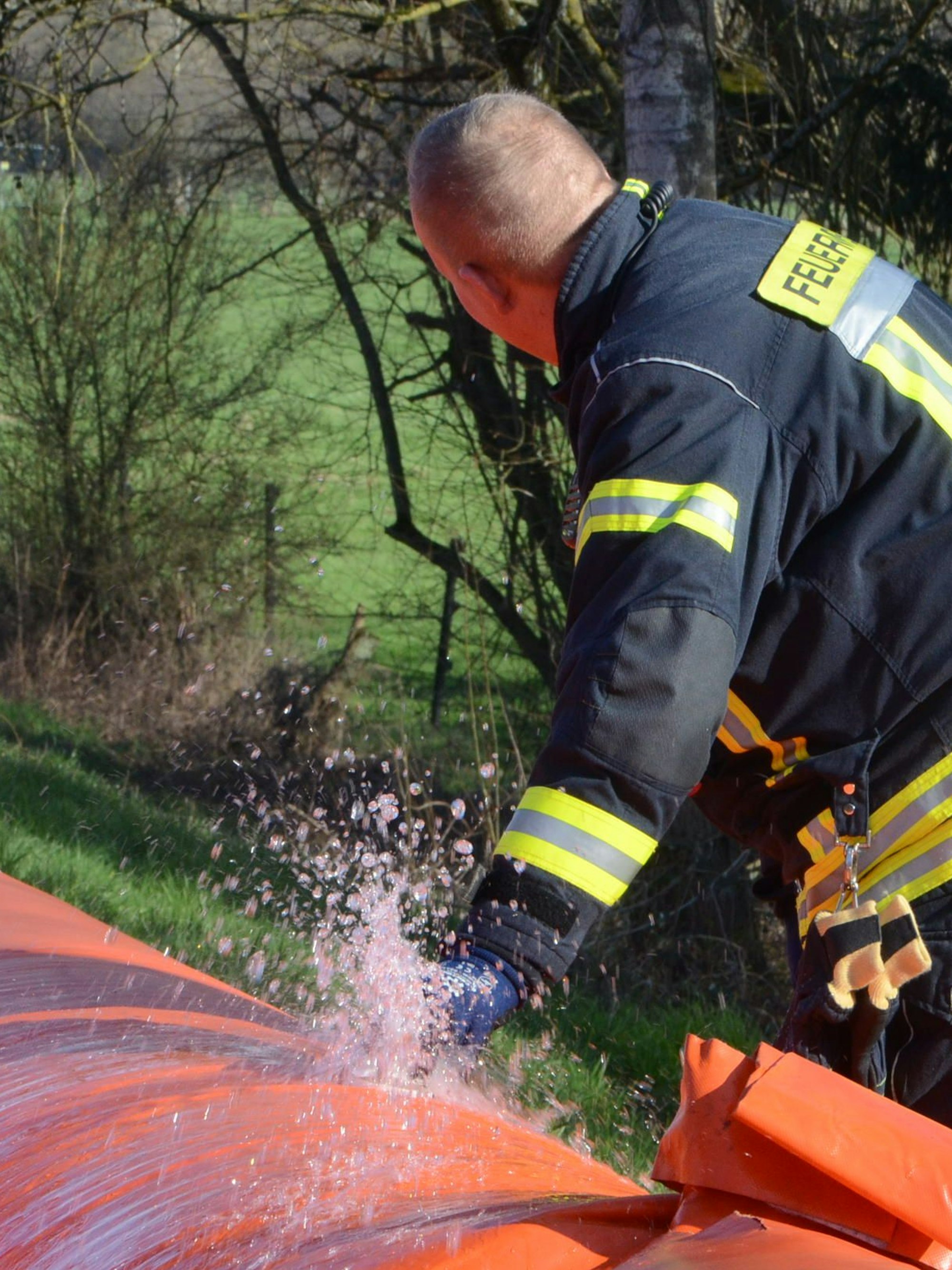 Ein Feuerwehrmann neben einem orange Kunststoffschlauch. Unter seiner Hand spritzt Wasser hervor.