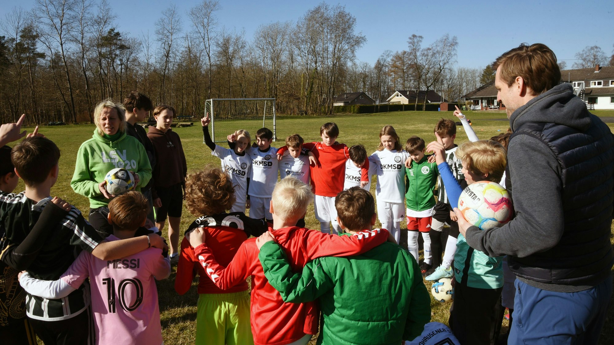 Eine Gruppe Kinder und zwei Trainer stehen in einem Kreis auf einem Fußballplatz zusammen.