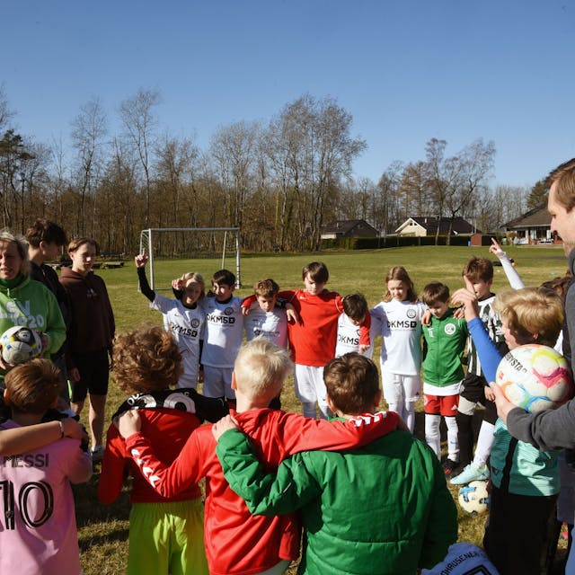 Eine Gruppe Kinder und zwei Trainer stehen in einem Kreis auf einem Fußballplatz zusammen.