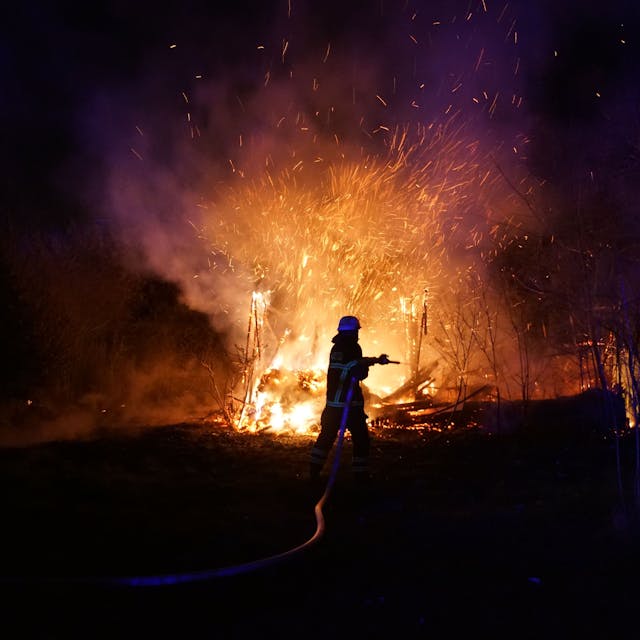 Eine Einsatzkraft der Feuerwehr steht mit einem Strahlrohr vor einem brennenden Schuppen. Funken schlagen in den Nachthimmel.