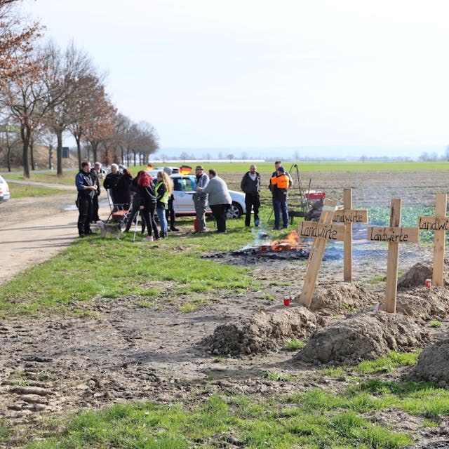 Eine Gruppe Menschen steht an einem Mahnfeuer auf einem Feld. Im Vordergrund sind symbolische Gräber zu sehen, die im Rahmen der Bauernproteste angelegt wurden.