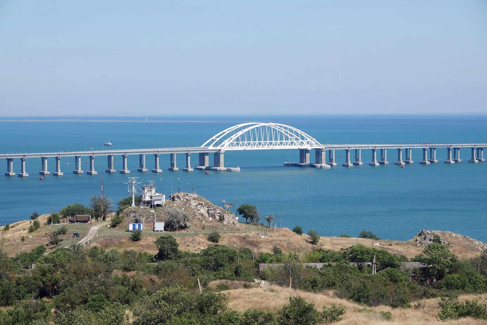 Eine Brücke führt über das Meer, im Vordergrund ist Land mit Sträuchern zu sehen.