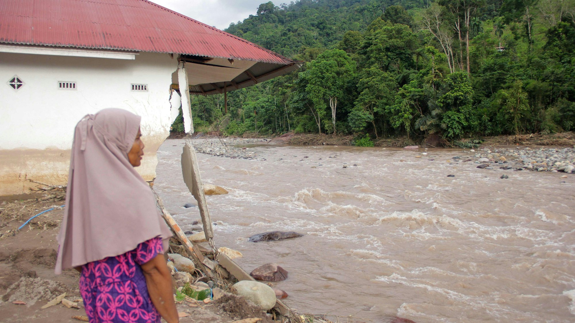 Eine Frau steht bei einem beschädigten Haus, von dem ein überfluteter Fluss große Stücke herausgebrochen hat.