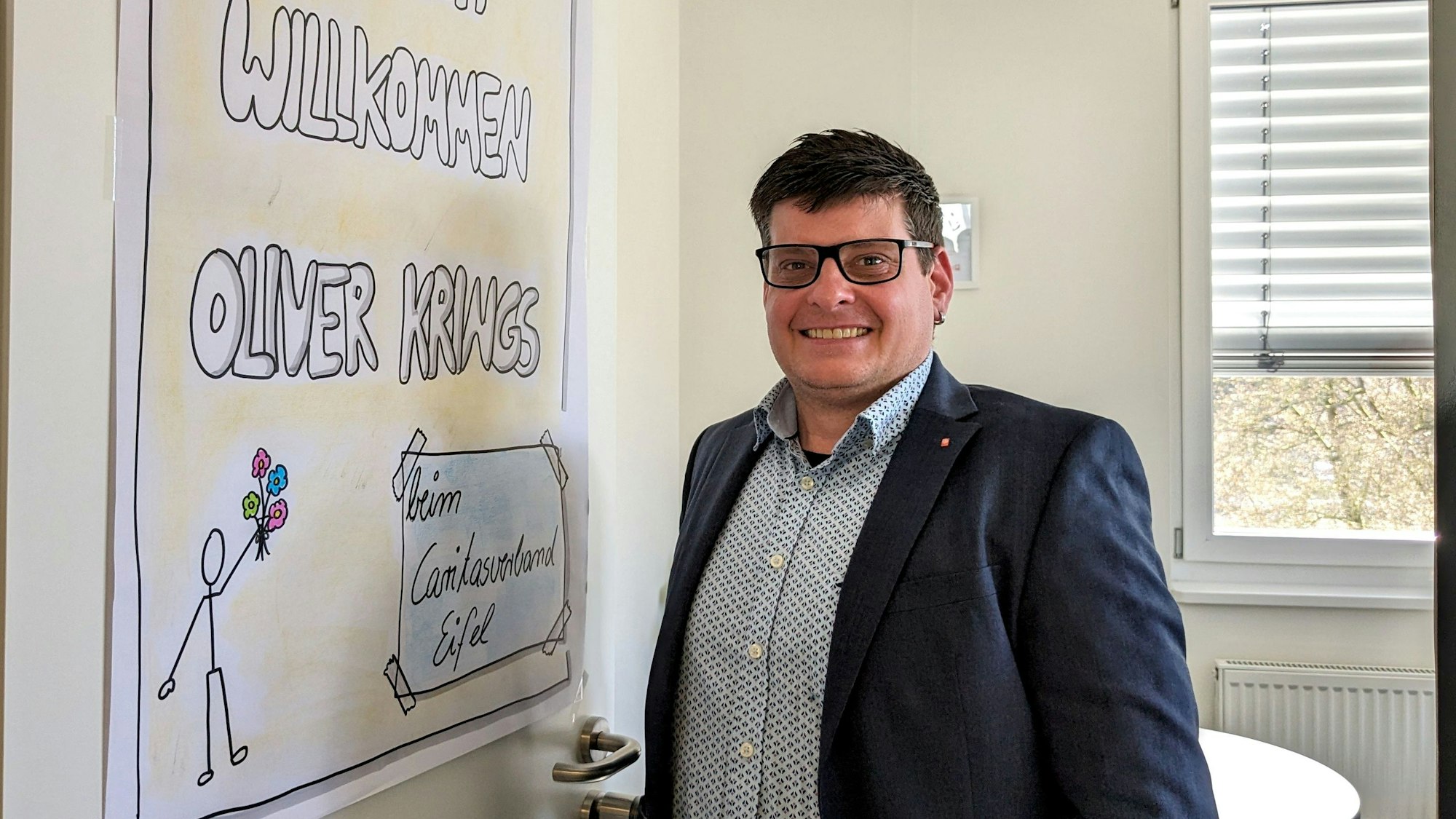 Oliver Krings steht an der Tür seines Büros, an dem ein Plakat mit der Aufschrift „Herzlich willkommen Oliver Krings“ zu sehen ist.