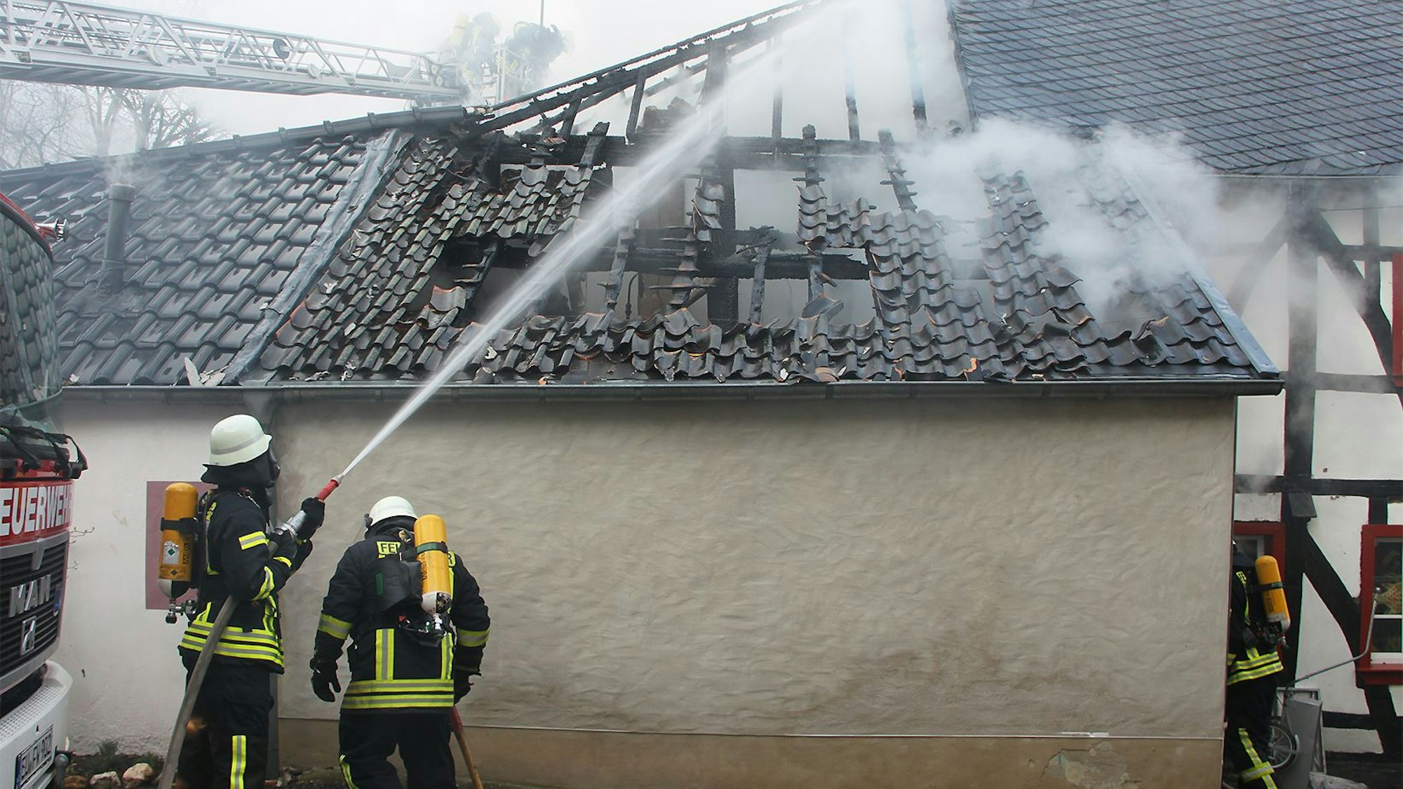 Zwei mit Atemschutzgeräten ausgerüstete Feuerwehrleute stehen an einem Gebäude und löschen einen Dachstuhl, der durch einen Brand schon massiv beschädigt ist.