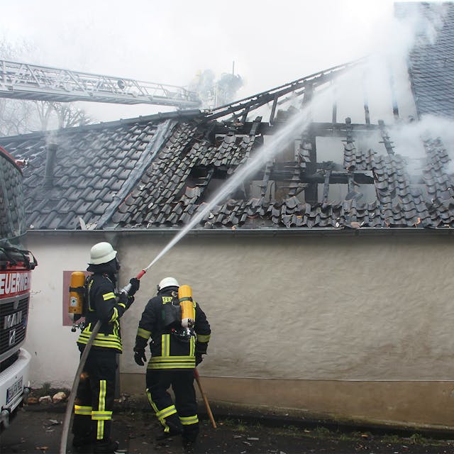 Zwei mit Atemschutzgeräten ausgerüstete Feuerwehrleute stehen an einem Gebäude und löschen einen Dachstuhl, der durch einen Brand schon massiv beschädigt ist.