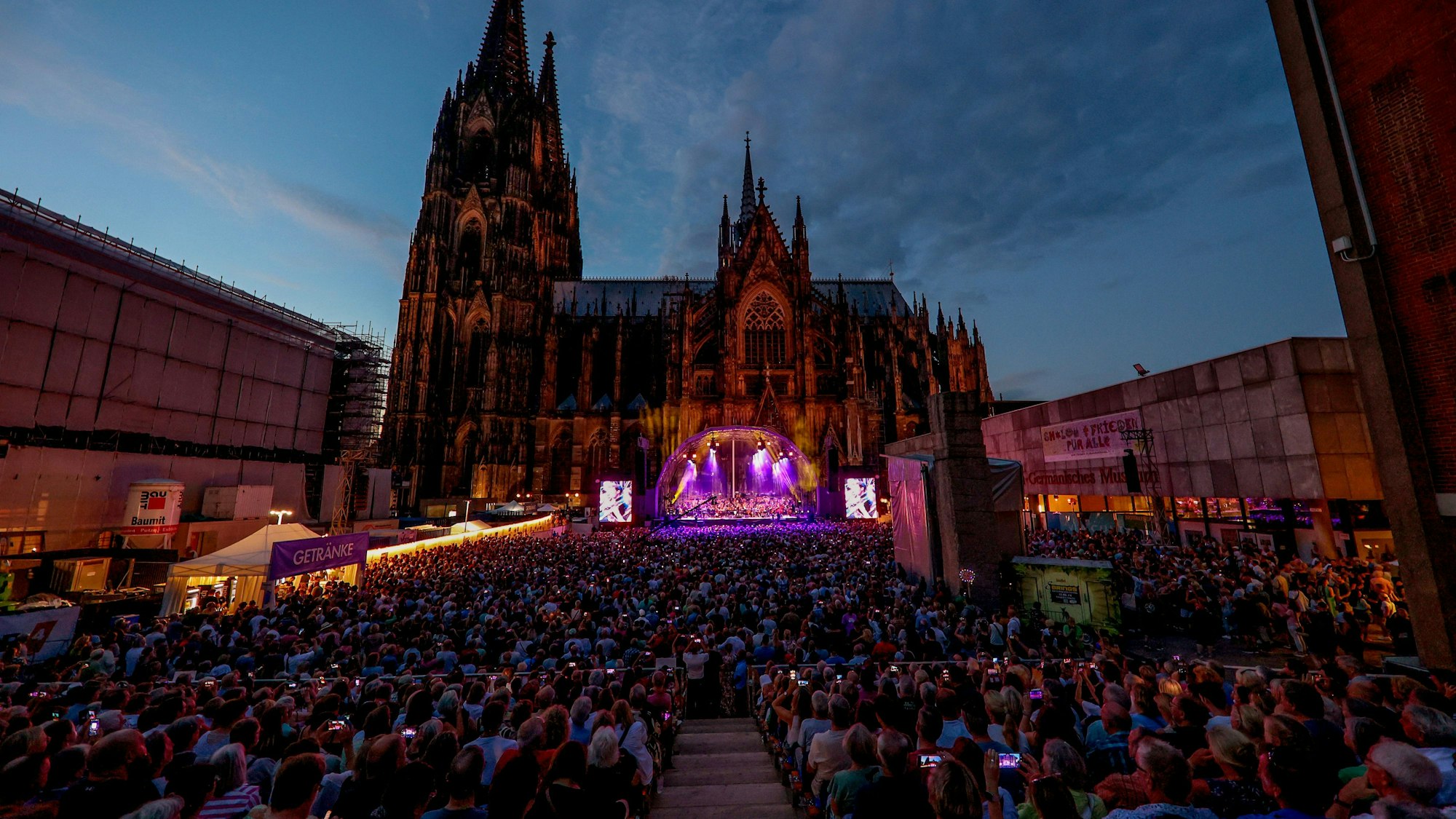 Zahlreiche Zuschauer sitzen in Reihen vor dem Kölner Dom, um auf dem Roncalliplatz einem Konzert zu lauschen.