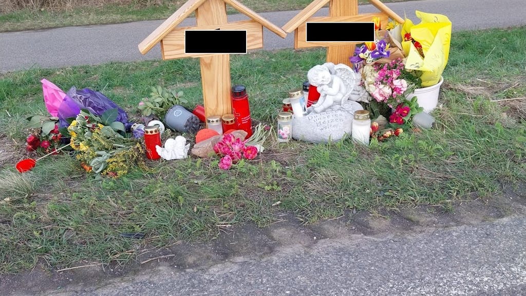 Zwei Holzkreuze stehen am Fahrbahnrand, davor Blumen, Kerzen und ein Engel aus Stein.