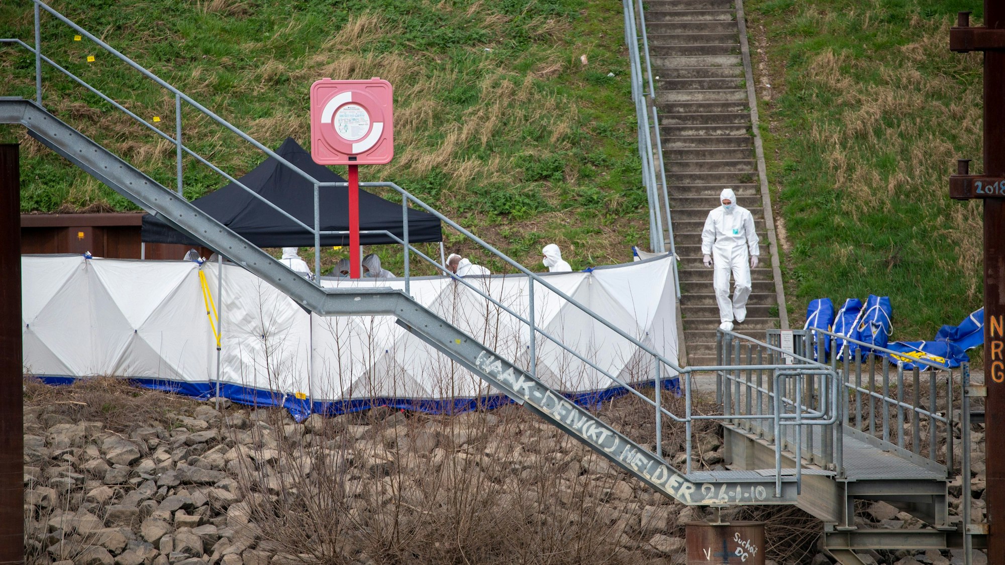 Hinter einem Sichtschutz sicherten Mitarbeitende der „Kriminaltechnischen Untersuchung“ am Sonntag Spuren im Mülheimer Hafen. Zu sehen sind Männer in weißen Schutzanzügen.