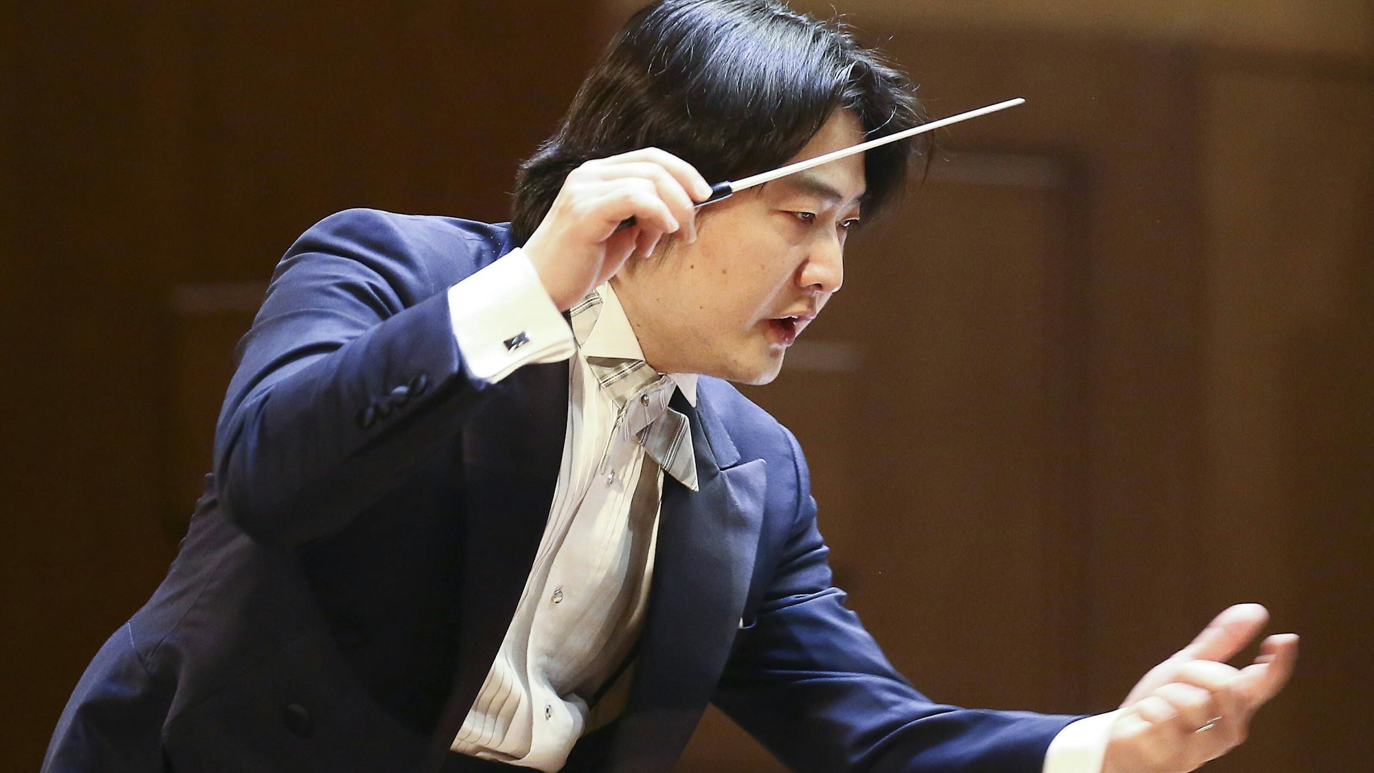 Auf dem Bild ist der Dirigent zu sehen. Er trägt einen blauen Anzug und ein weißes Hemd. Er hält in der rechten Hand seinen Taktstock und gestikuliert auch mit der Linken.