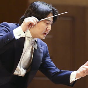 Auf dem Bild ist der Dirigent zu sehen. Er trägt einen blauen Anzug und ein weißes Hemd. Er hält in der rechten Hand seinen Taktstock und gestikuliert auch mit der Linken.&nbsp;