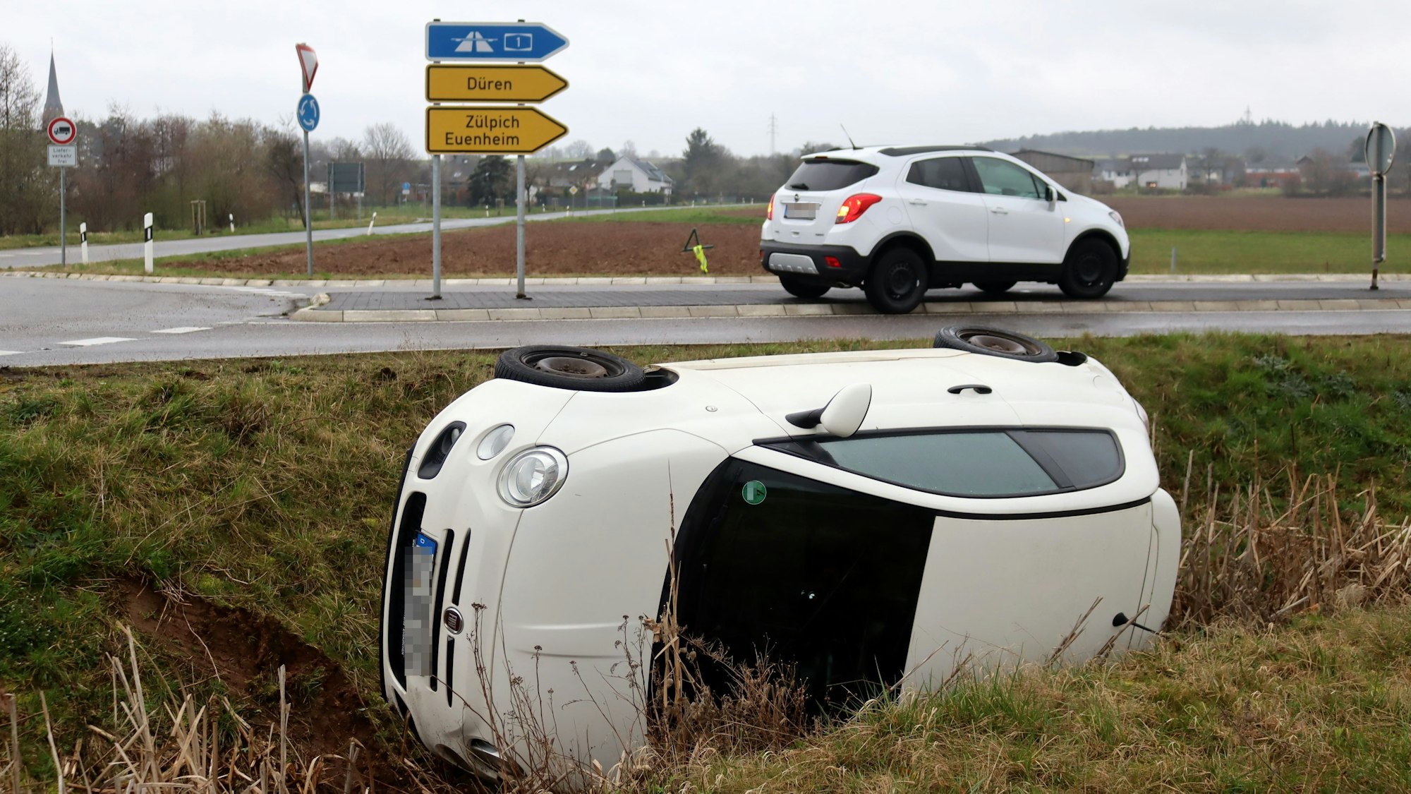Ein weißer Fiat 500 liegt nach einem Unfall bei Billig im Straßengraben neben einem Kreisverkehr.