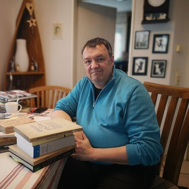 Autor Ralf Thiesen an seinem Arbeitsplatz in seinem Haus. Sein Tisch ist voll beladen mit Fachbüchern, die er für die Recherche seines Romans genutzt hat.