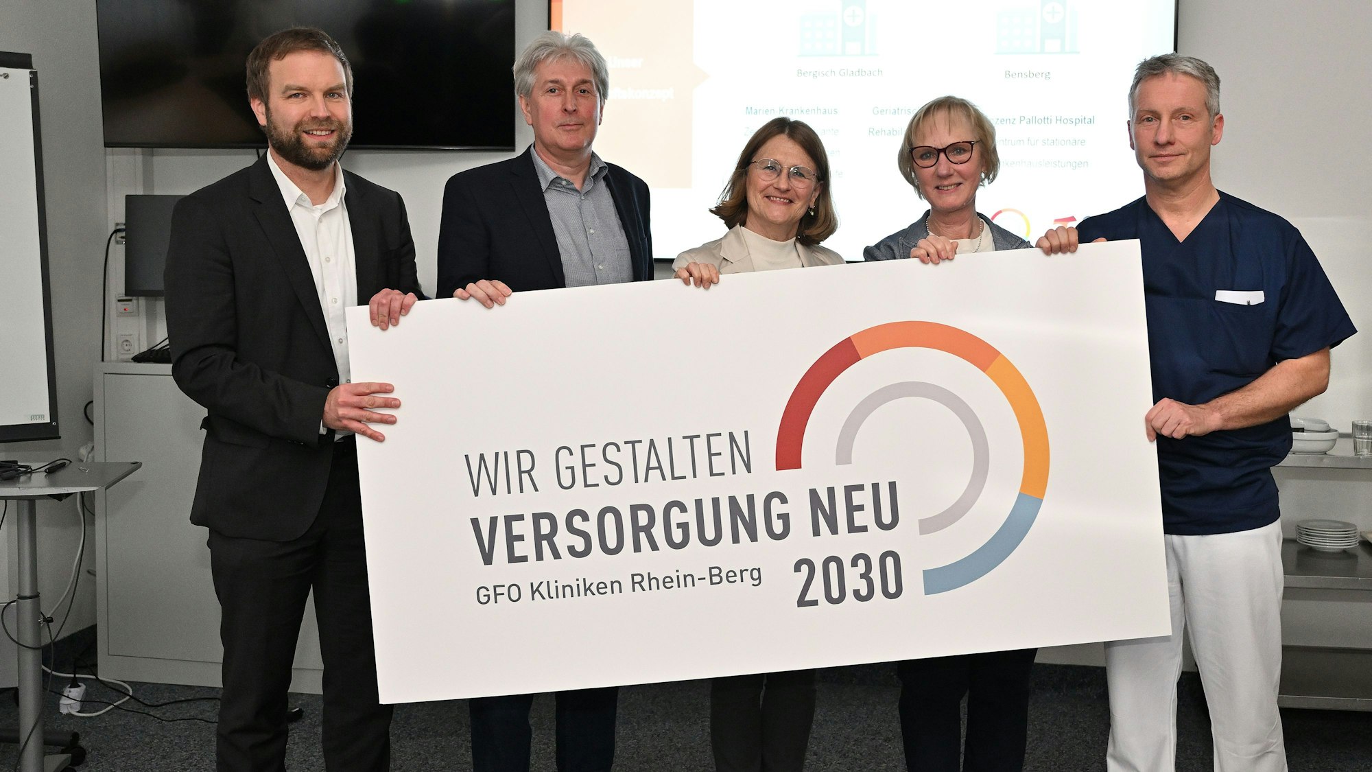 Stephan Muhl, Dr. Jörg Kösters, Dr. Barbara Florange, Monika Gerfer und Dr. Stefan Machtens halten ein Schild, auf dem steht: „Wir gestalten Versorgung neu. GFIO Kliniken Rhein-Berg 2030“.