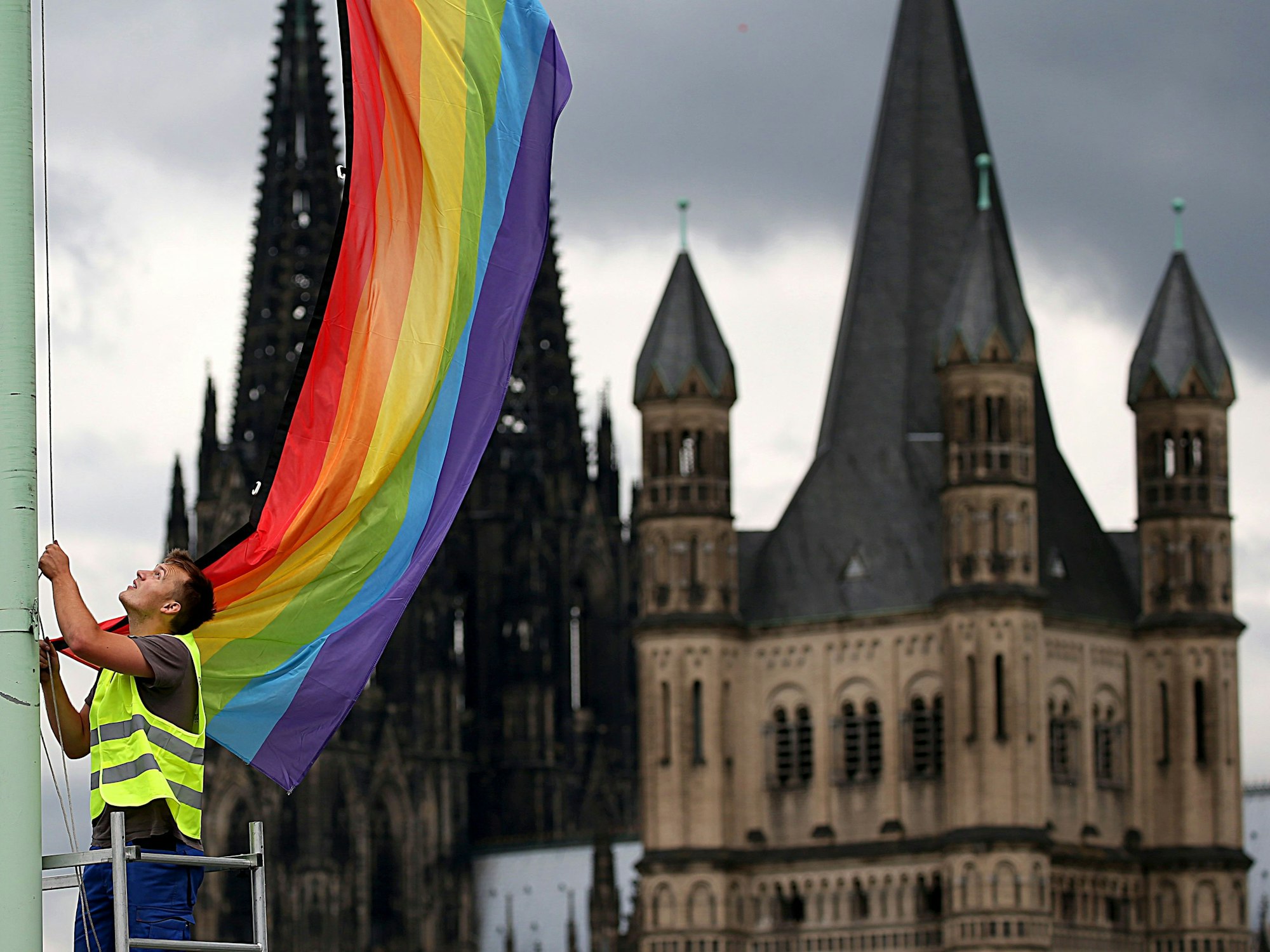 Ein Arbeiter hängt eine Regenbogenfahne vor der Kulisse des Doms und der Kirche Groß St. Martin auf.