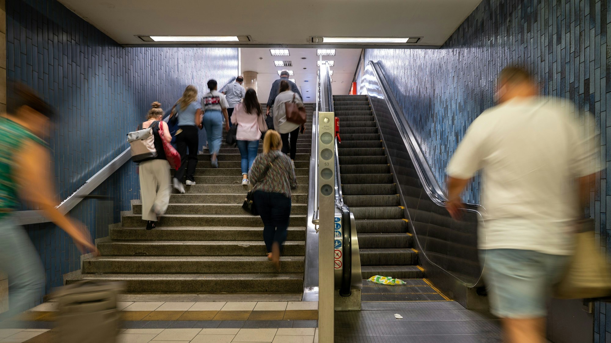 Menschen steigen eine Treppe an der KVB-Haltestelle Dom/Hauptbahnhof hoch, ein Mann geht auf die Rolltreppe zu.