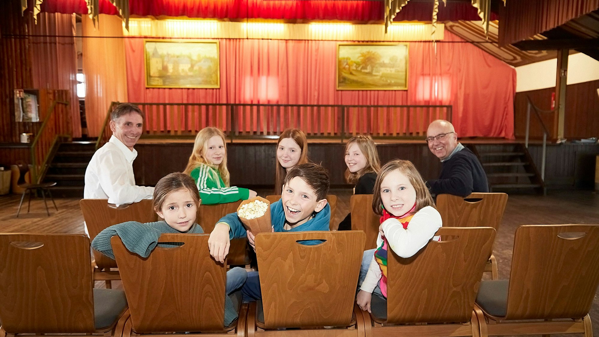 Sechs Kinder und zwei Herren haben auf den Stühlen im Dorfsaal Platz genommen. Ein Junge hält eine Tüte mit Popcorn in der Hand.