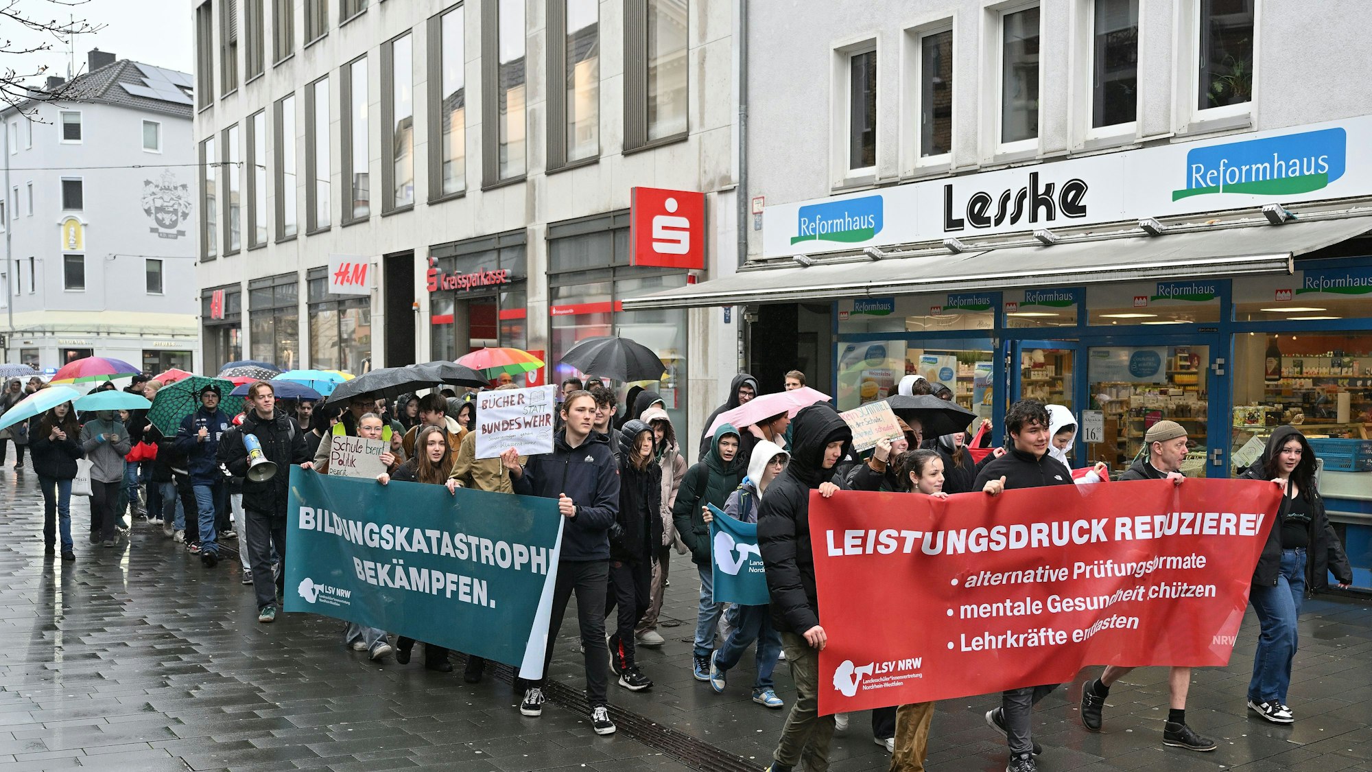 Vorneweg gehen Schüler und tragen ein Banner. Es folgen weitere Demonstranten, teils mit Plakaten, teils mit Regenschirmen in der Hand. Im Hintergrund sieht man die Fassaden von Geschäften.