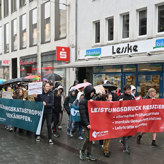 Vorneweg gehen Schüler und tragen ein Banner. Es folgen weitere Demonstranten, teils mit Plakaten, teils mit Regenschirmen in der Hand. Im Hintergrund sieht man die Fassaden von Geschäften.&nbsp;