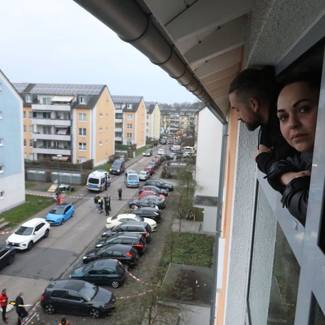 Eine Frau schaut aus einem Fenster auf den abgesperrten Tatort vor einem Wohnhaus in Vingst.
