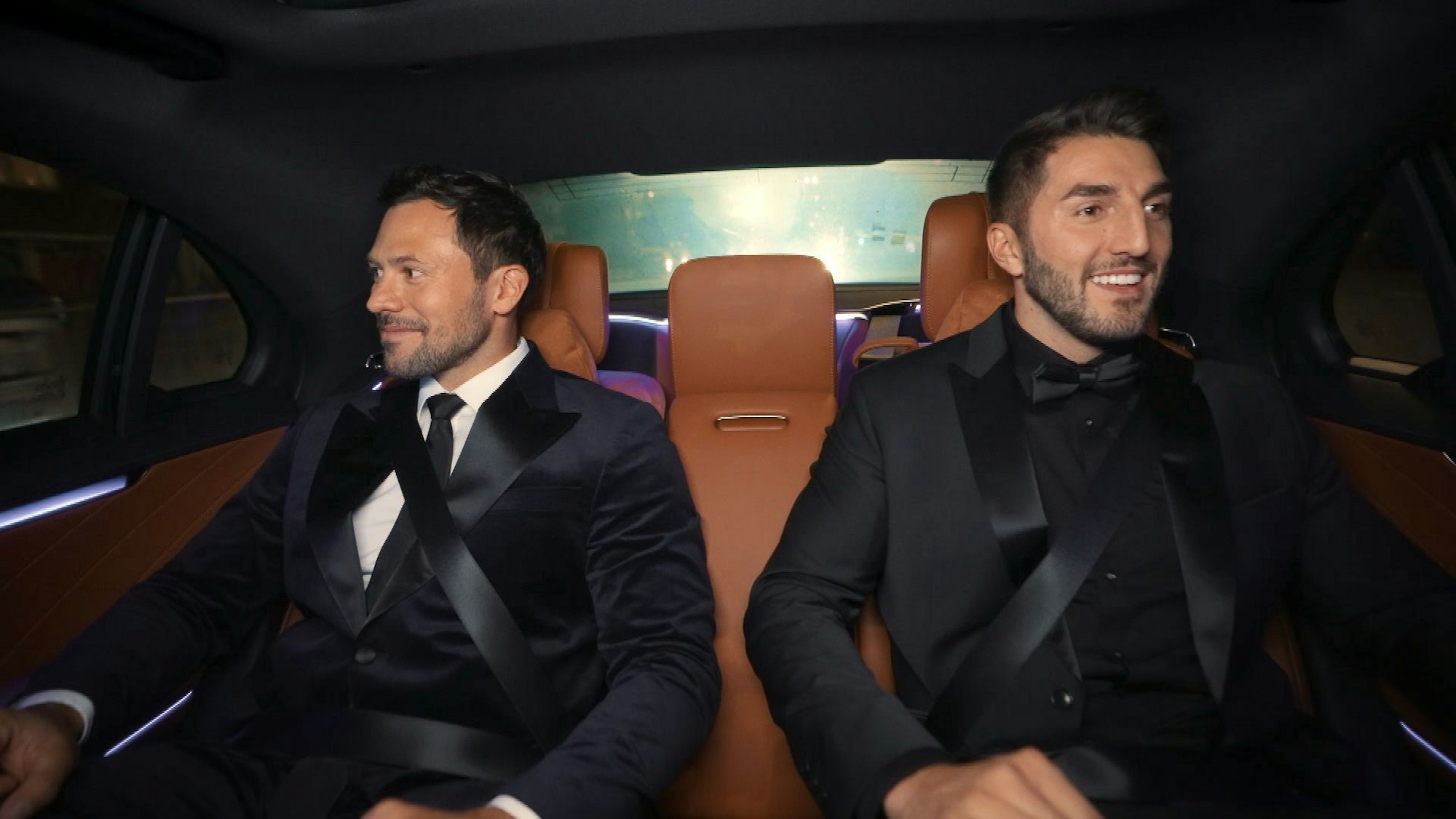 Zwei Männer in Anzügen sitzen in einem Auto nebeneinander.