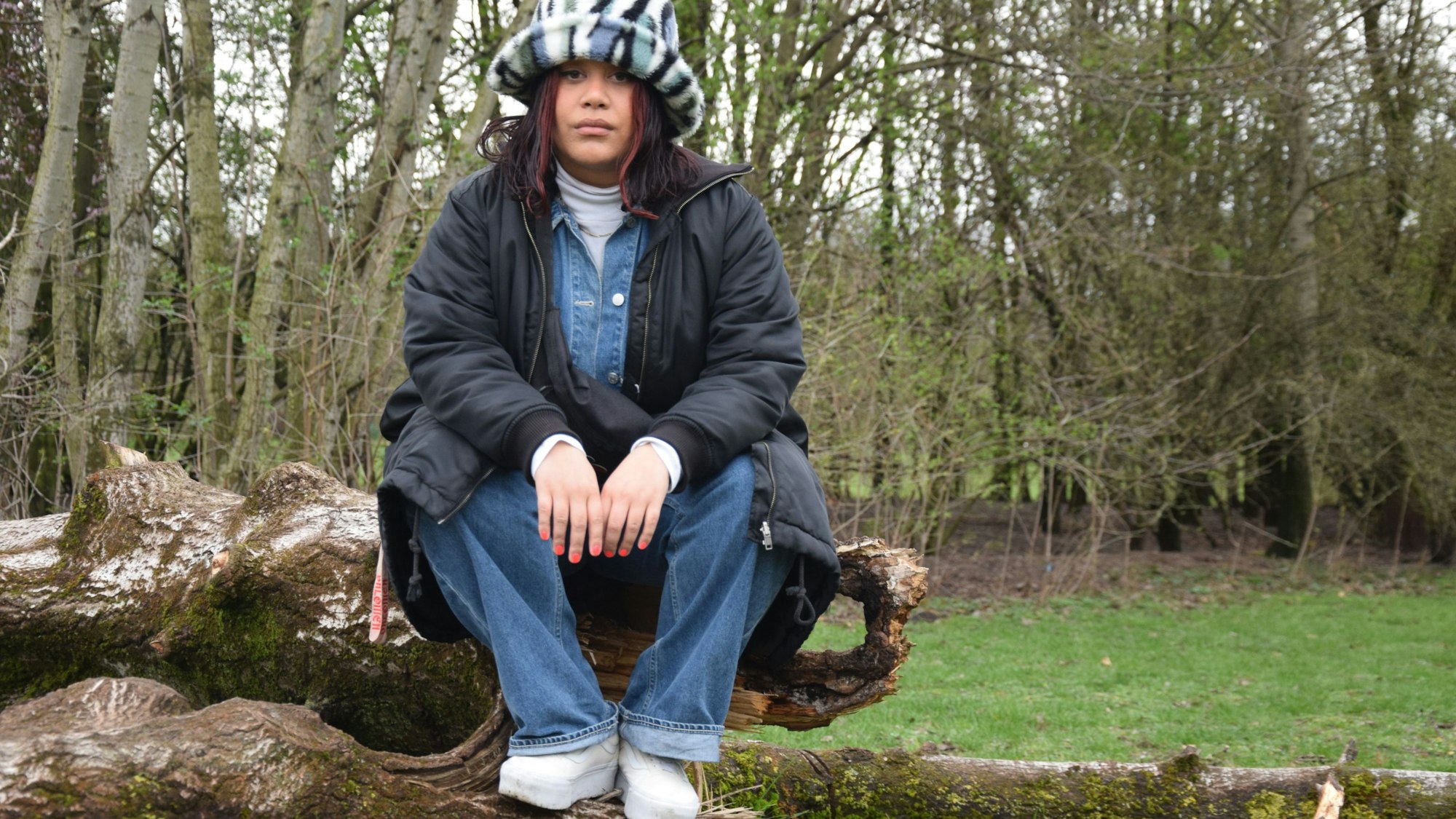 Eine junge Frau sitzt in Hut und Jacke auf einem Baumstammstapel.