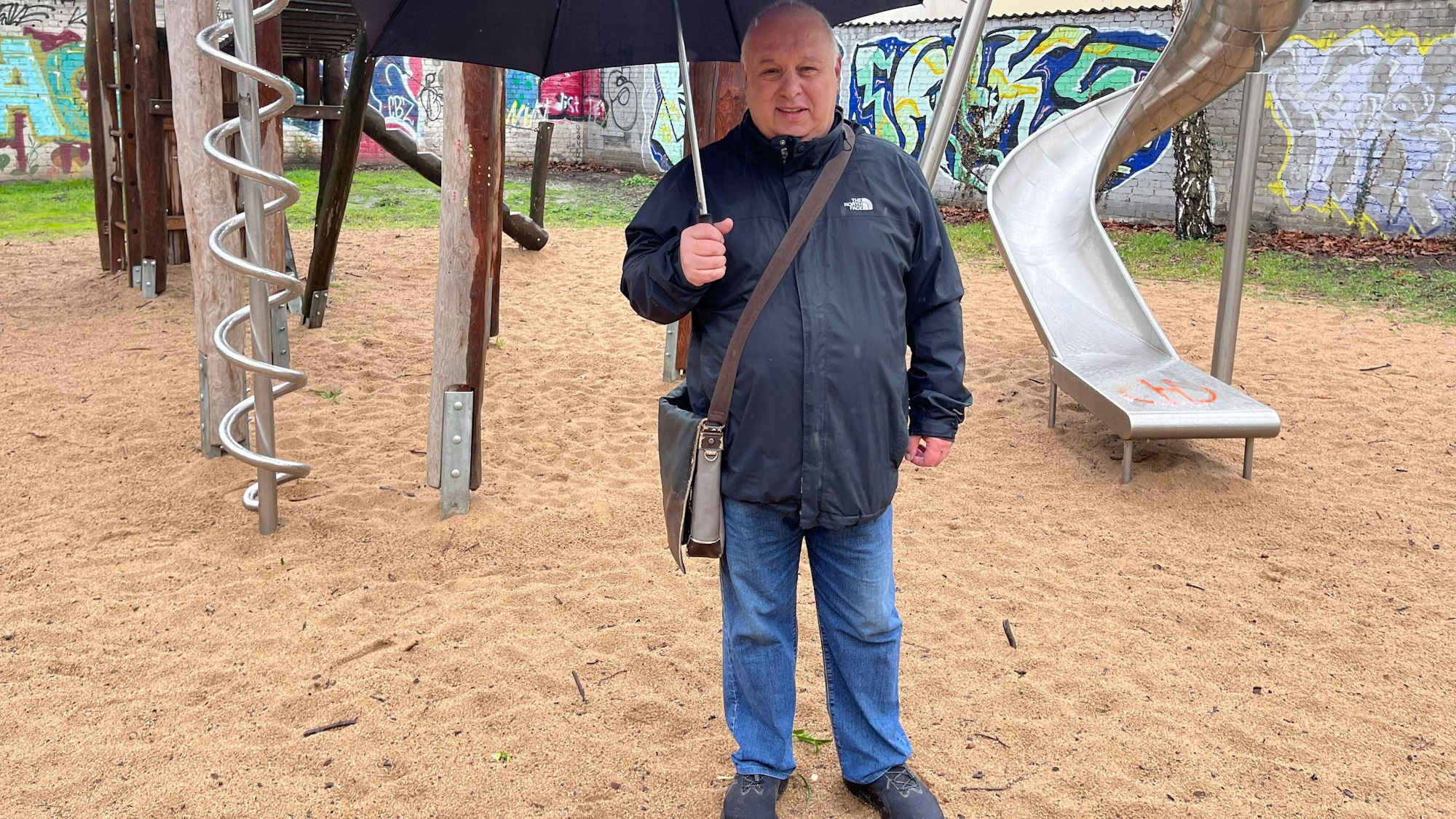 Ein Mann mit Regenschirm steht vor einer Kletter-Rutsche-Kombination auf einem Spielplatz.