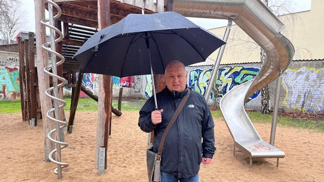 Ein Mann mit Regenschirm steht vor einer Kletter-Rutsche-Kombination auf einem Spielplatz.