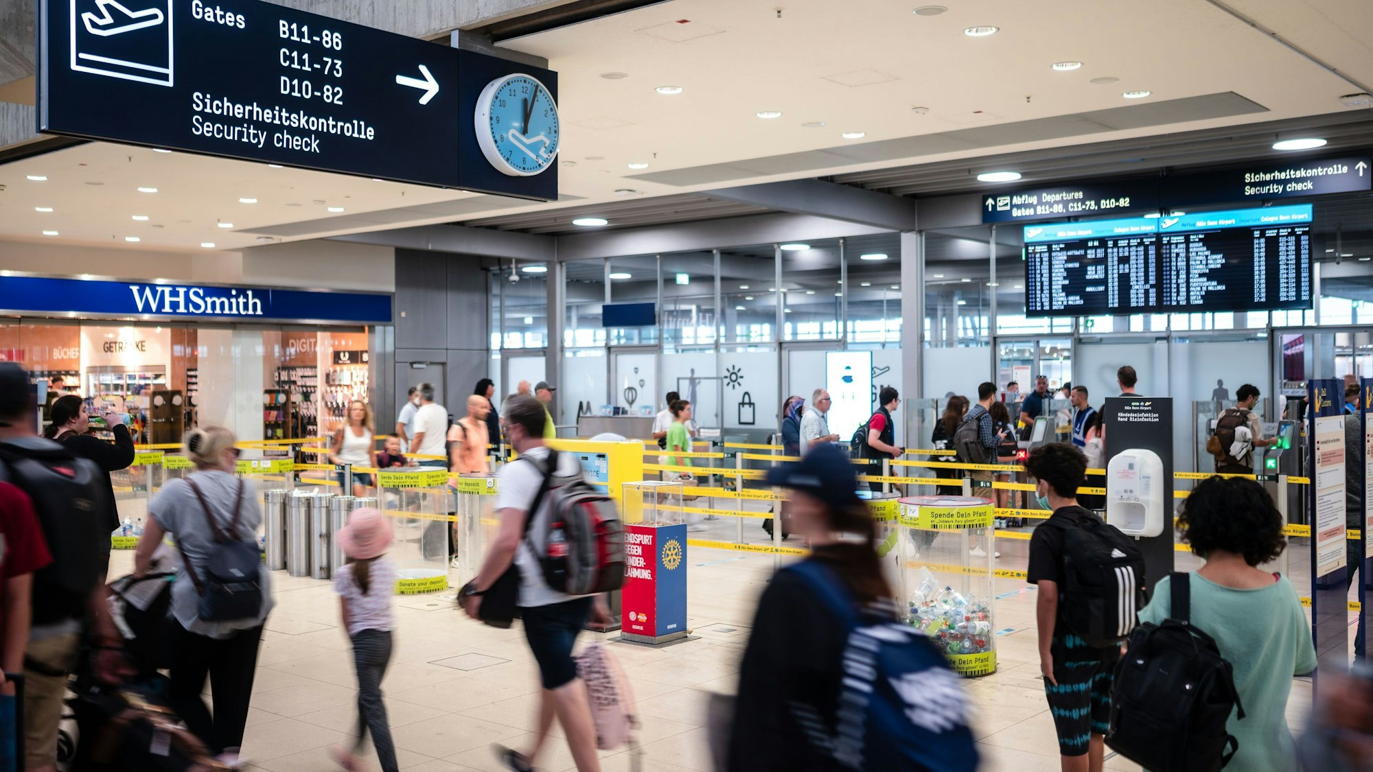 Menschen gehen am Flughafen Köln/Bonn durch die Hallen, im Hintergrund sind die Sicherheitskontrollen zu erkennen.