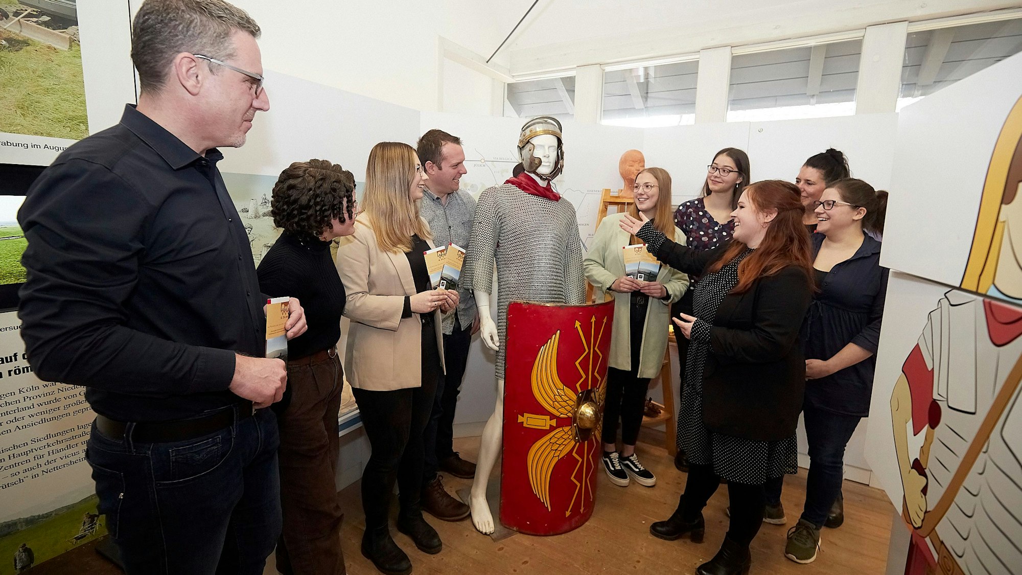 Eine Gruppe Männer und Frauen steht in Nettersheim in einem Ausstellungsraum an einer Figur, die eine nachgebaute Rüstung eines römischen Soldaten trägt.
